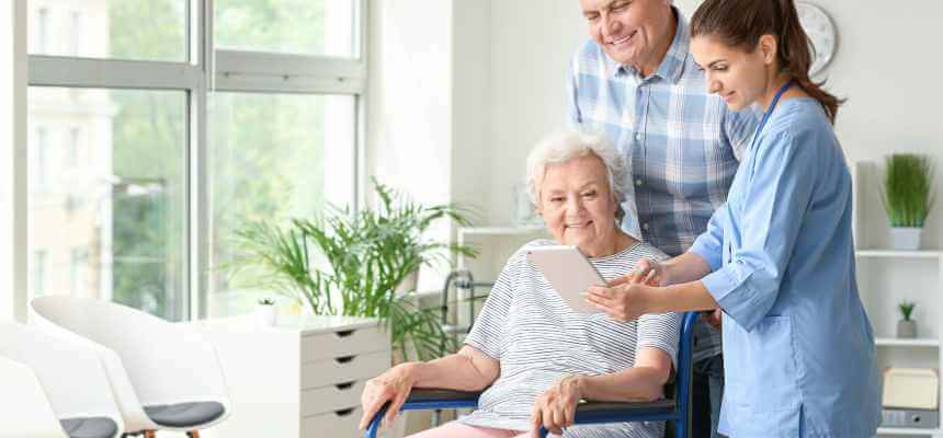Ældre dame sidder smilende i stol med en plejehjemsansat, der viser hende en tablet. En pårørende står bag kvinden.